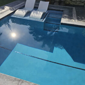 Section of pool in a walled enclosure, finished with Blue Topaz by Luxury Pools of Miami. The pool is accented by blue glass tiles and has a sun shelf with two loungers.