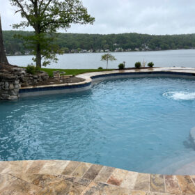 A serene freeform pool overlooking a river with brown travertine pavers and coping for a deck. There is a large tree on the upper left of the image, surrounded by a field stone wall. The pool was finished with Diamond Brite French Gray by All State Technologies, Inc.