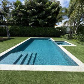 Relaxing backyard pool area with a cabana striped awning on the left of the image and beautifully landscaped yard in the background. The pool itself is has light gray travertine coping around the pool and spa. There is no deck, the pool is surrounded by manicured green grass. The pool itself is a rectangular shape with the sunken spa centered on the right side. Subway tile in varying shades of teal surround the waterline and the pool was finished by Pugh's Pools in Diamond Brite Watercolors Isla Verde.