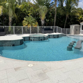 Beautiful light gray stone pool deck on two levels surrounds a freeform pool with small glass tile in shades of black, gray and white. The lower pool has a gray marble counter area on the edge of a sun shelf area with matching swim up stools located next to it. The upper deck area has a spa with a water feature cascading into the pool. There are seating areas on the upper deck, with a small shed and gazebo on the lower deck. Diamond Brite Blue Quartz was applied by Seagull Backyards.